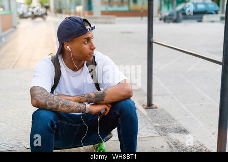 Tatuato giovane uomo seduto sul suo skateboard ascoltando musica con lo smartphone e auricolari Foto Stock