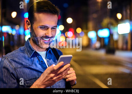 Uomo sorridente con cuffie wireless utilizza lo smartphone nella città di notte Foto Stock