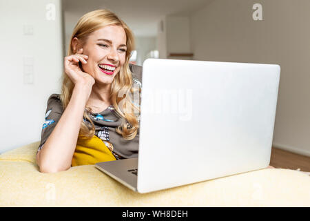 Ridendo giovane donna utilizzando laptop sul lettino a casa Foto Stock