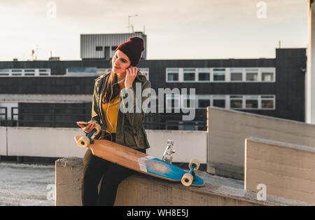 Elegante giovane donna con lo skateboard, gli auricolari e il telefono cellulare sul ponte di parcheggio Foto Stock