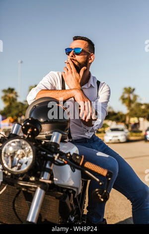 Ritratto di barbuto motociclista in mirroring con occhiali da sole seduto su una moto di fumare Foto Stock