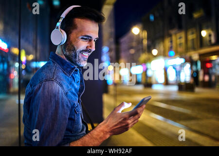 Uomo sorridente con cuffie utilizza lo smartphone mentre si è in attesa per il bus notturno in città Foto Stock