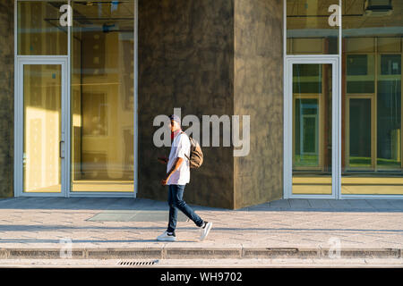 Giovane uomo con zaino camminando sul marciapiede alla luce del sole Foto Stock