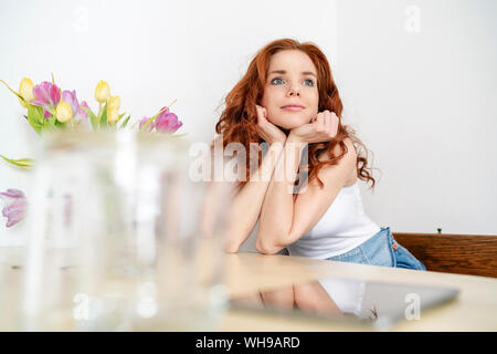 Riflessivo redhead metà donna adulta seduto con le mani sul mento mentre appoggiata sul tavolo a casa Foto Stock