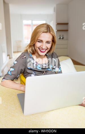 Ridendo giovane donna utilizzando laptop sul lettino a casa Foto Stock