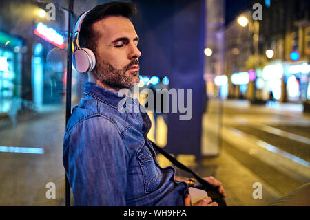 Uomo con cuffie wireless snoozing durante l'attesa per il bus notturno in città Foto Stock