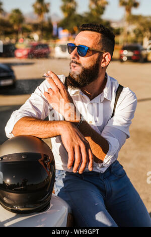 Ritratto di barbuto motociclista in mirroring con occhiali da sole appoggiano sul suo casco di fumare Foto Stock