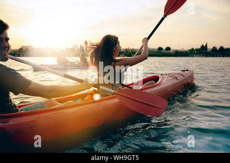 Fiducioso giovane coppia caucasica kayak sul fiume insieme con il tramonto del sole negli sfondi. Divertimento in attività del tempo libero. Romantica e felice donna e uomo in kayak. Sport, concetto di relazioni. Foto Stock