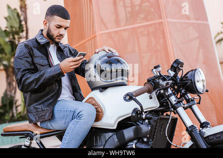 Giovane uomo seduto sulla sua moto tramite telefono cellulare Foto Stock
