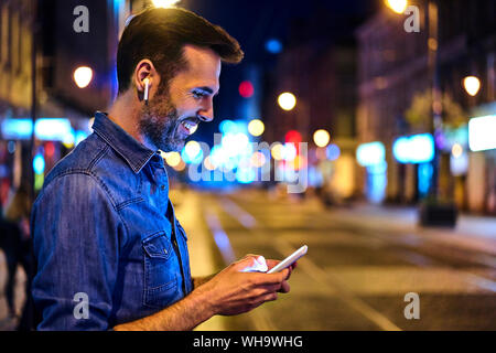 Uomo con cuffie wireless utilizza lo smartphone nella città di notte Foto Stock