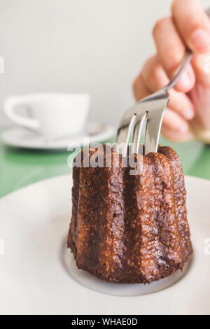 Traveler donna mano che tiene una forcella e taglio di un Canelé (Canele - Caneles de Bordeaux) tradizionale francese dessert dolci da forno bianco con tazza di caffè b Foto Stock