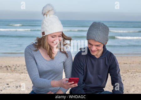 Felice maschio e femmina modelli indossano berretti a maglia con altoparlanti Bluetooth interno, ascoltando musica insieme con mobile smart phone. Guardando alla m. Foto Stock