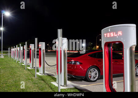 Stazione Supercharger Tesla di notte con Tesla Model S collegata e in carica in background. Foto Stock