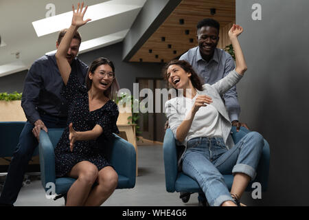 Carefree multirazziale imprenditori divertendosi in ufficio a cavallo su sedie Foto Stock