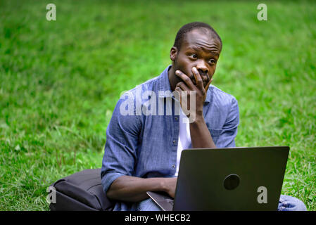 Perplesso studente africano lad da portatile su un parco Prato Foto Stock