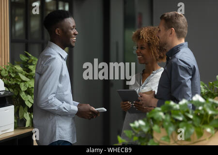 Tre diversi colleghi felici parlando di ridere in piedi in ufficio Foto Stock