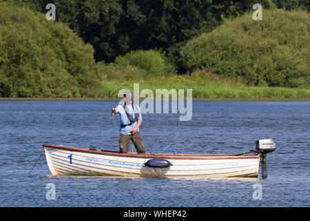 Ravensthorpe Reservoir, Northamptonshire, Regno Unito: Un pescatore con un cappuccio piatto si trova in una piccola barca con un motore fuoribordo e getta la sua linea. Foto Stock