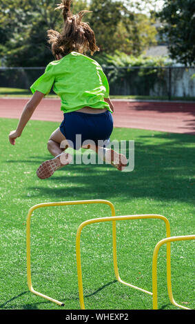 Alta scuola ragazza sta saltando su un piede di due mini giallo hurdle suddivisione i suoi piedi a lato durante la via pratica. Foto Stock