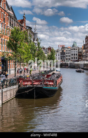 Amsterdam, Paesi Bassi - 1 Luglio 2019: Dark flat intrusione viene utilizzato come galleggianti parcheggio bici sul canale Singel sotto blu-grigio cloudscape. Torre Munt spi Foto Stock