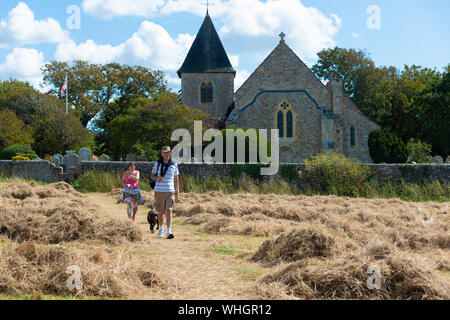 Dog walkers passeggiando attraverso un hayfield davanti alla chiesa normanna di San Pietro e di San Paolo a West Wittering,West Sussex, in Inghilterra, Regno Unito Foto Stock