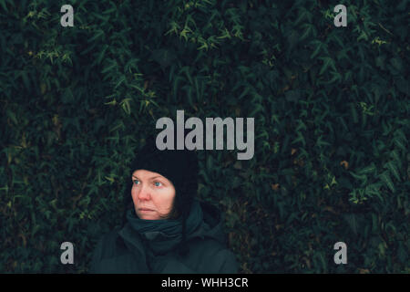 Grave donna in abiti invernali contro la parete di edera om fredda giornata invernale, guardando triste e preoccupato Foto Stock