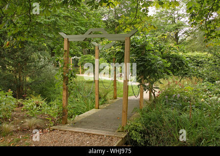 Un piccolo ponte con un arco in legno si attraversa un piccolo ruscello in un tranquillo giardino inglese Foto Stock