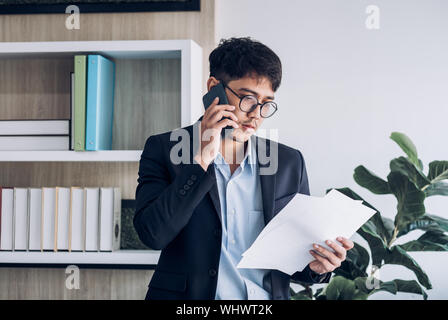 Giovane imprenditore asiatici indossare tuta parlando al cellulare e vedere la documentazione permanente al tavolo scrivania in ufficio moderno.Il contatto con il concetto del cliente Foto Stock