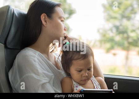 Bambina è la lettura di un libro sulla sua madre giro mentre sua madre esausta sguardi fuori dalla finestra Foto Stock