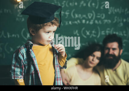 Smart bambino nel cappuccio laureati da eseguire. Ragazzo presentando la sua conoscenza a mamma e papà. I genitori in ascolto il loro figlio, lavagna su sfondo, defoc Foto Stock