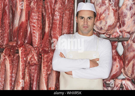 Ritratto di un macellaio in piedi con le braccia incrociate davanti la carne cruda Foto Stock