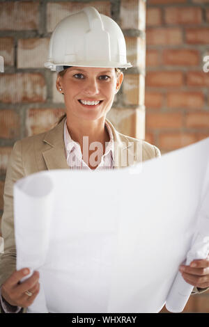 Ritratto di una donna sorridente architetto in hardhat con blueprint al sito in costruzione Foto Stock