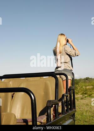 Vista posteriore di una giovane donna bionda sui piedi di safari in jeep cerca attraverso il binocolo Foto Stock