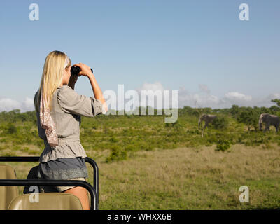 Vista posteriore di una giovane donna bionda sui piedi di safari in jeep cerca attraverso il binocolo Foto Stock