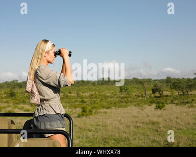 Vista laterale di una giovane donna bionda sui piedi di safari in jeep cerca attraverso il binocolo Foto Stock