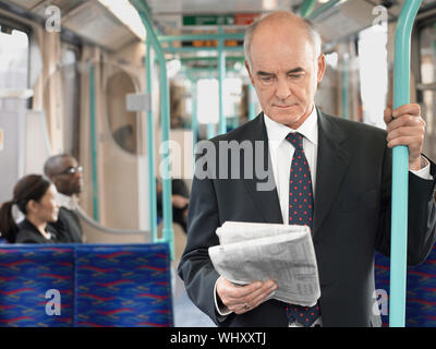 Imprenditore maturo leggendo il giornale in treno con i pendolari seduti in background Foto Stock