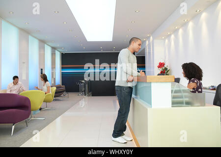 Uomo che parla di receptionist presso il banco del ricevimento in ufficio Foto Stock