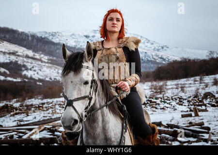 Bella ragazza a cavallo e con i capelli rossi in armatura. La donna è un Vichingo. Fantasy Foto Stock