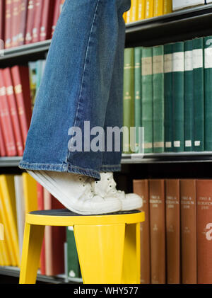 Closeup lowsection di un giovane uomo in piedi su uno sgabello per raggiungere libro in biblioteca Foto Stock