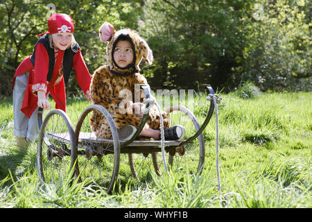 Giovane ragazzo in costume pirata spingendo pensieroso ragazzo in jaguar costume sul carrello in giardino Foto Stock