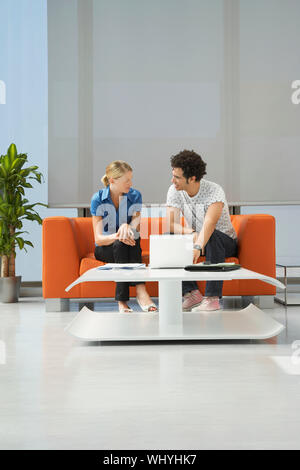Per tutta la lunghezza di una coppia multietnica tramite computer portatile sul divano arancio in sala di ricevimento presso l'ufficio Foto Stock