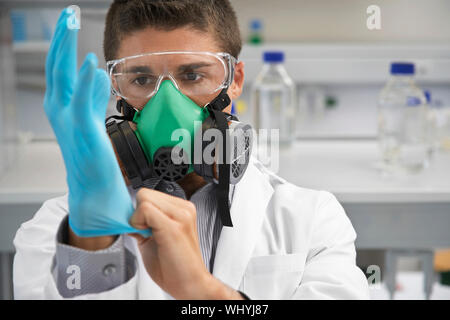 Scienziato maschio in una maschera a gas mentre mettendo sul guanto di gomma in laboratorio Foto Stock