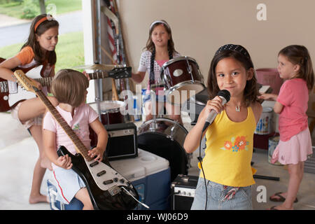 Ritratto di giovane carina ragazza cantare nel microfono con gli amici suonare uno strumento musicale in garage Foto Stock