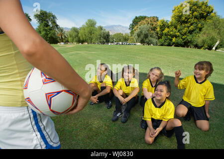 Pullman in piedi nella parte anteriore del gruppo della ragazza i giocatori di calcio (7-9 anni) seduto sul prato, vista in elevazione Foto Stock