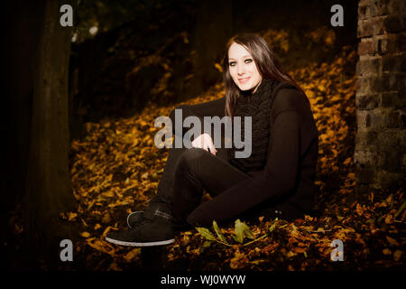 Bella donna di foglie di autunno seduto fuori nelle tenebre sul terreno a fianco di un muro di mattoni, con copyspace Foto Stock