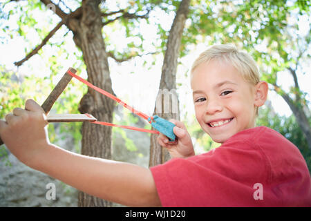 Vista laterale verticale di una bionda boy utilizzando slingshot contro tronchi di alberi Foto Stock