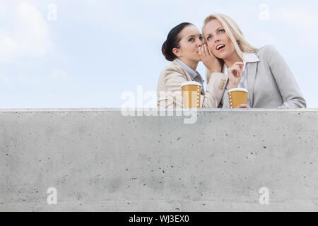 Basso angolo vista di imprenditrice whispering in collega l orecchio mentre si sta in piedi sul terrazzo contro sky Foto Stock
