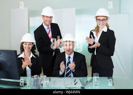 Team di quattro diversi architetti o ingegneri strutturali hardhats indossa in un ufficio ad applaudire Foto Stock