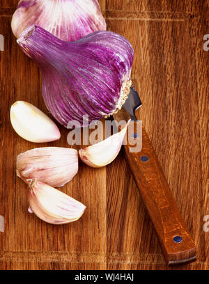 Disposizione dei freschi aglio rosa corpo pieno e fette con coltello da cucina sul tagliere di legno closeup Foto Stock