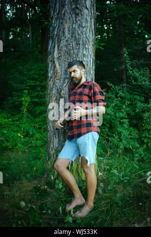 Traveler in plaid shirt e Jean Corti il viaggio a piedi nudi. Uomo Barbuto con pala in foresta. Hipster con barba lunga sul naturale paesaggio verde Foto Stock