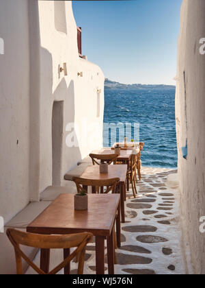 Brown tavoli e sedie di street ristorante collocato nel passaggio stretto tra l'edificio nei pressi di increspatura mare sull'isola di Mykonos in Grecia Foto Stock